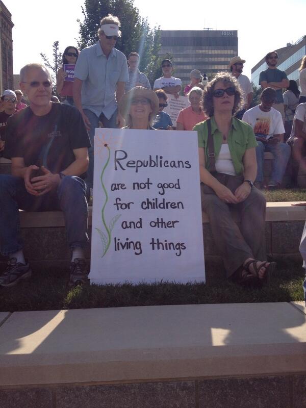A man and woman sitting beside a woman holding a sign that says Republicans are not good for children and other living things